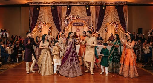 Family members dancing together under warm Sangeet stage lights